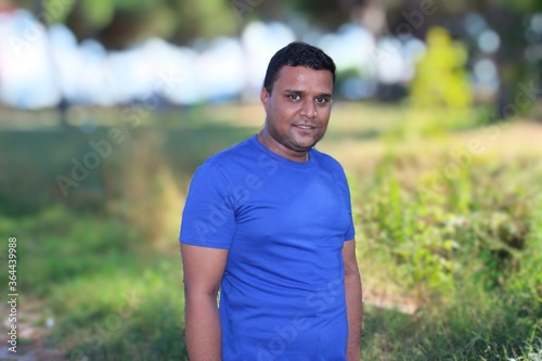 close portrait of young man in blue t shirt standing outside in park with trees in background © Ali