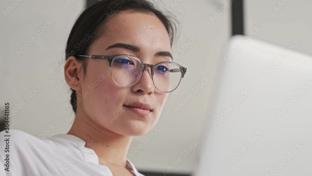 Close up view of Calm elegant pretty asian woman in eyeglasses using laptop computer while sitting on sofa at home Close up view of Calm elegant pretty asian woman in eyeglasses using laptop computer while sitting on sofa at home
