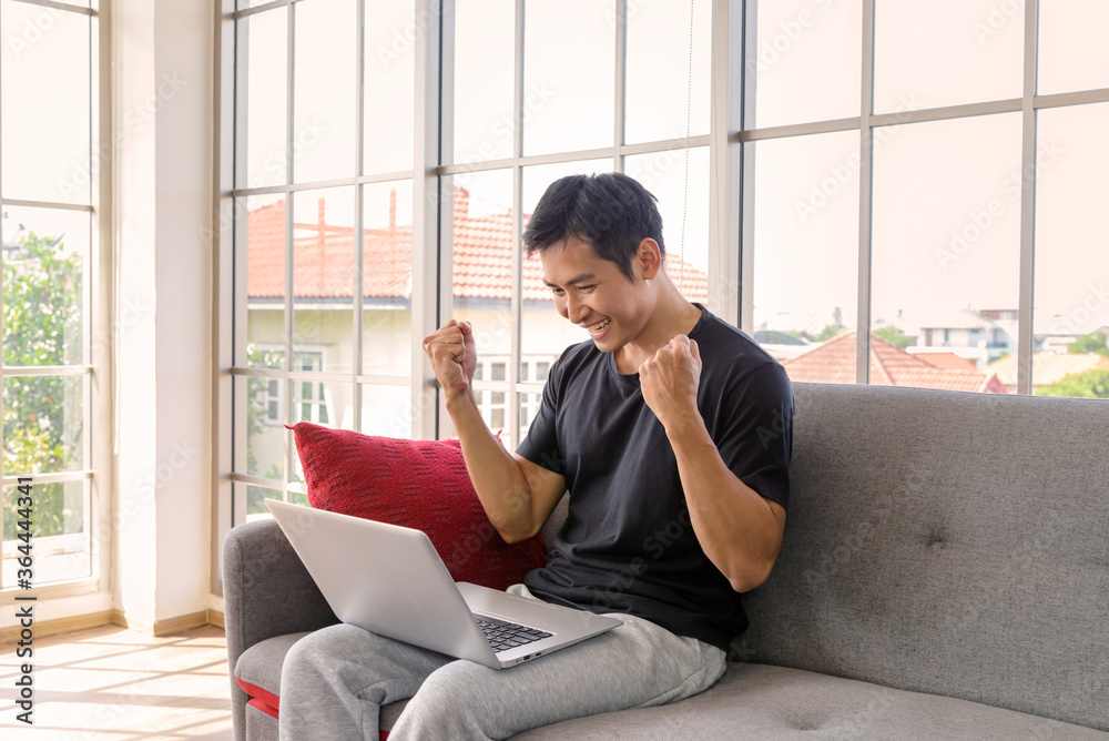 Happy asian man sitting on cozy couch. his punching the air while working with laptop computer. successful concept