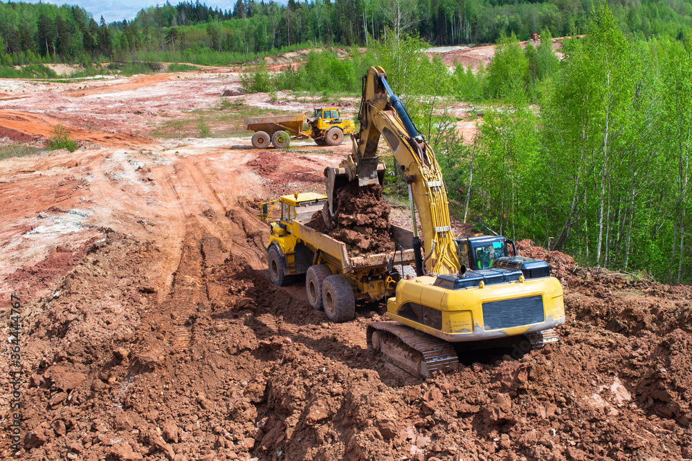 Clay mining. The yellow excavator digs red clay with a large bucket and puts it into the truck. Clay mining. The yellow excavator digs red clay with a large bucket and puts it into the truck.
