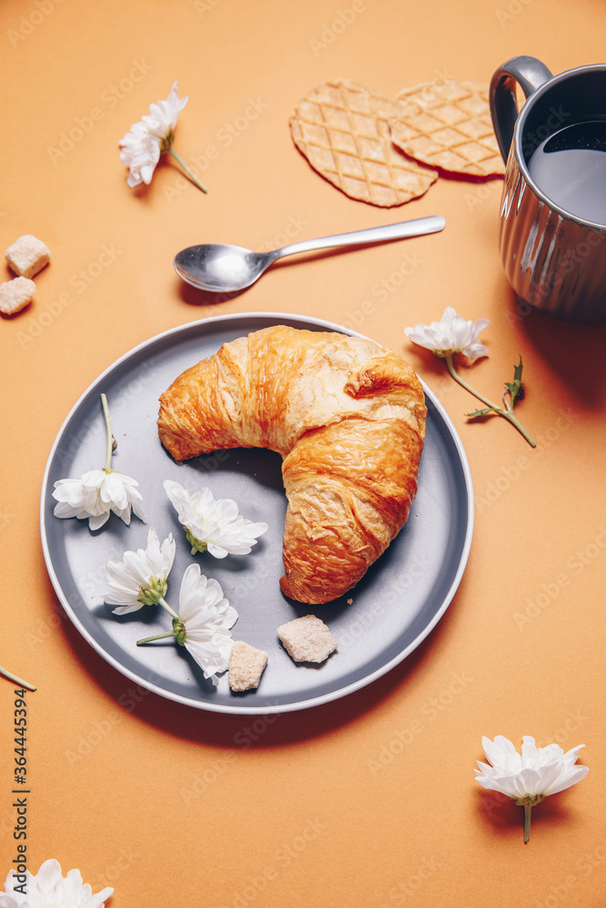 croissant with small white chrysanthemum flowers on an orange background. Breakfast with tea, teaspoon and croissant on an orange background.