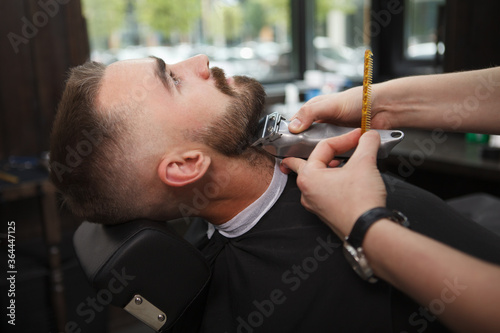 Male client relaxing while professional barber trimming his beard