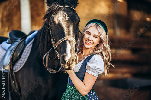 Pretty blonde in traditional dress take care of big black horse at the farm