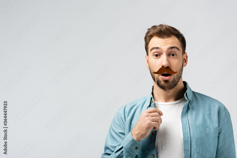 shocked young man with fake mustache on stick isolated on grey