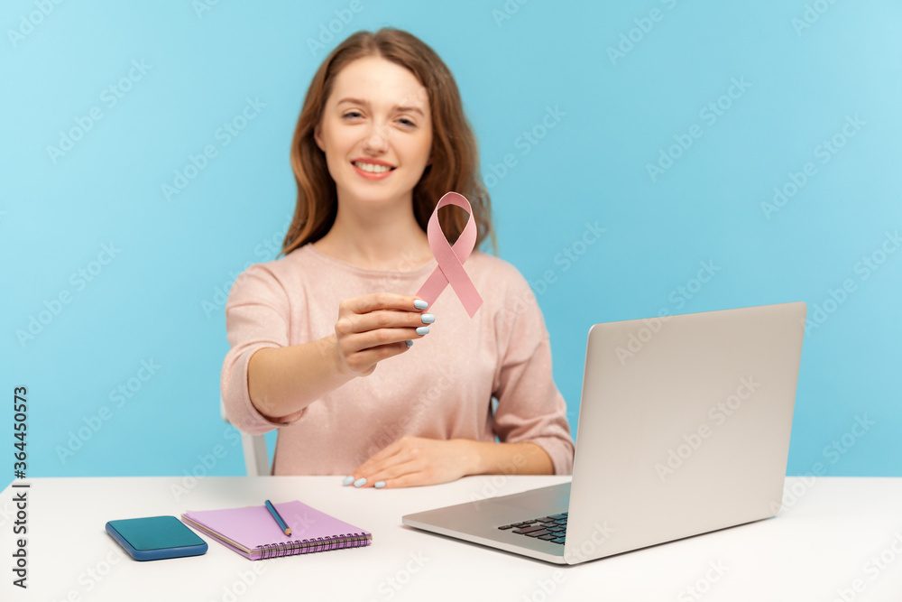 Optimistic pretty woman, volunteer or manager, sitting at workplace with laptop and showing pink ribbon, symbol of breast cancer awareness, female health. indoor studio shot isolated, blue background Optimistic pretty woman, volunteer or manager, sitting at workplace with laptop and showing pink ribbon, symbol of breast cancer awareness, female health. indoor studio shot isolated, blue background