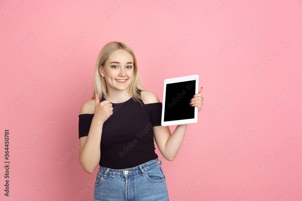 Showing tablet, thumb up. Portrait of young woman with bright emotions on coral pink studio background. Blonde female model. Concept of human emotions, facial expression, sales, advertising, youth. Showing tablet, thumb up. Portrait of young woman with bright emotions on coral pink studio background. Blonde female model. Concept of human emotions, facial expression, sales, advertising, youth.
