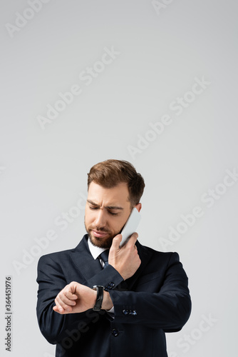 serious handsome businessman in suit talking on smartphone and looking at wristwatch isolated on grey