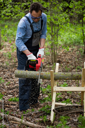 Close up view of woodcutter lumberman in working uniform sawing small tree trunk on sawhorse with electric saw chainsaw in forest on sawmill, outdoors. Wood work, handwork, wood cutting tools, timber