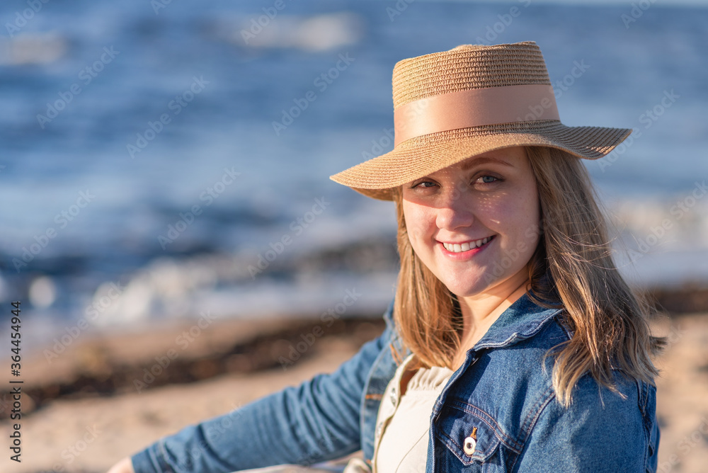 The concept of peace of mind and happiness. A woman with blond hair in a straw hat and a denim jacket stands on the background of the sea and enjoys the sun.