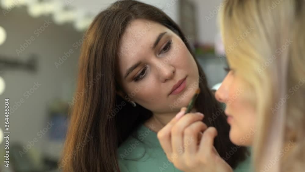 Side view of professional make-up artist applying eyeshadow to face of beautiful young woman in beauty salon, close-up. Concept of beauty, fashion and stylish makeup. Shooting in slow motion. Side view of professional make-up artist applying eyeshadow to face of beautiful young woman in beauty salon, close-up. Concept of beauty, fashion and stylish makeup. Shooting in slow motion.