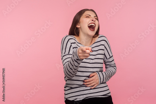 Ha ha, you're ridiculous! Portrait of woman in striped sweatshirt pointing fingers to camera and laughing out loud, teasing funny friend appearance. indoor studio shot isolated on pink background