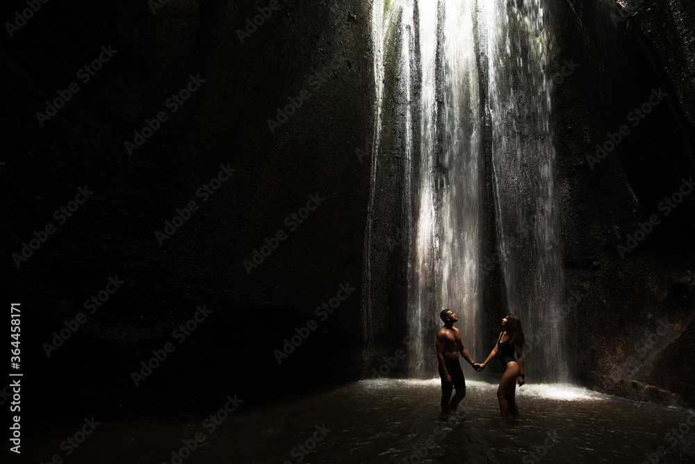 Beautiful couple in a cave with a waterfall. Athletic man and woman under the streams of a waterfall. A sexy couple under a tropical waterfall in twilight light. Tukad Chepung Waterfall. Copy space Beautiful couple in a cave with a waterfall. Athletic man and woman under the streams of a waterfall. A sexy couple under a tropical waterfall in twilight light. Tukad Chepung Waterfall. Copy space