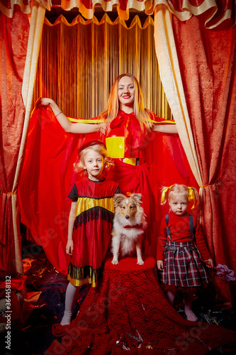 Family with dog during stylized theatrical circus photo shoot in beautiful red location. Model mother and daughters with small animal posing on stage with curtain