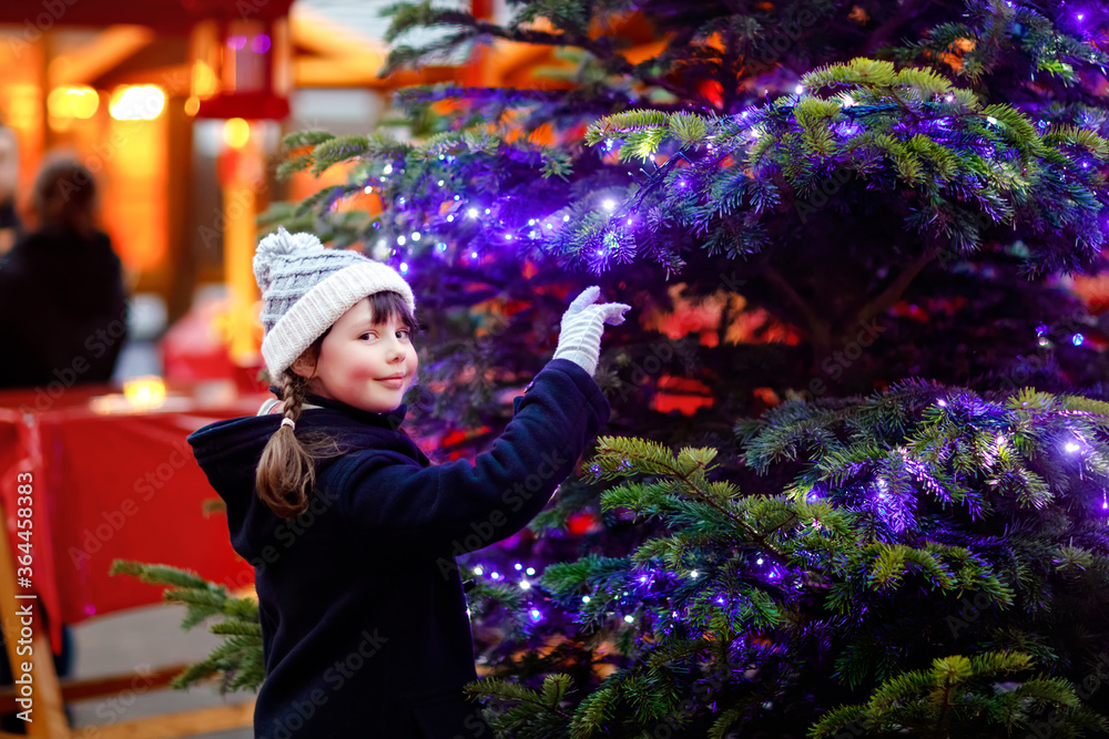 Little cute kid girl having fun on traditional Christmas market during strong snowfall. Happy child enjoying traditional family market in Germany. Schoolgirl standing by illuminated xmas tree.