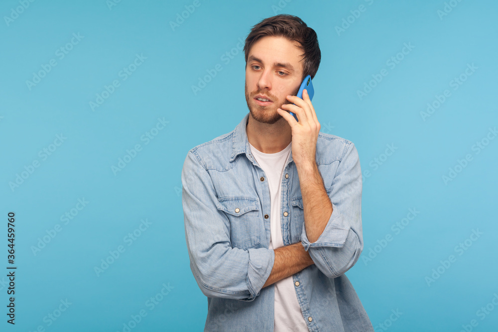 Portrait of young handsome businessman in casual denim shirt talking on phone, having serious mobile conversation with partner, making important call. indoor studio shot isolated on blue background Portrait of young handsome businessman in casual denim shirt talking on phone, having serious mobile conversation with partner, making important call. indoor studio shot isolated on blue background