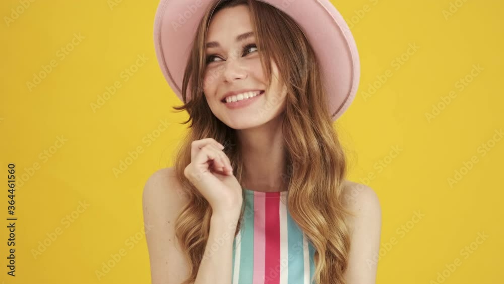 A joyful young woman in a pink hat is posing and hiding her face isolated over a yellow background A joyful young woman in a pink hat is posing and hiding her face isolated over a yellow background