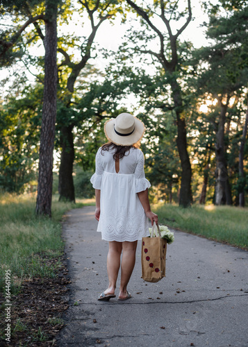 A beautiful girl in a white dress and a straw hat with hydrangea in a basket walks through the forest. Self-isolation. unity with nature © Julia