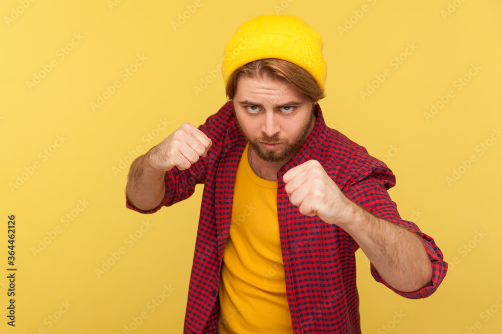 Aggressive hipster bearded guy in beanie hat, checkered shirt holding clenched fists up ready to boxing, punching with confident expression, fighting spirit. studio shot isolated on yellow background