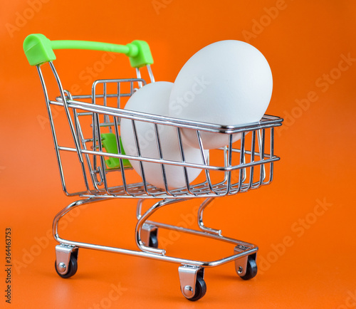 Two eggs in a small shopping cart. Ingredient for Easter cake.  Mini trolley with eggs on a orange background. Selective focus. Copy space. photo