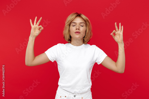 Calm young blonde woman in casual white outfit meditating over red background.