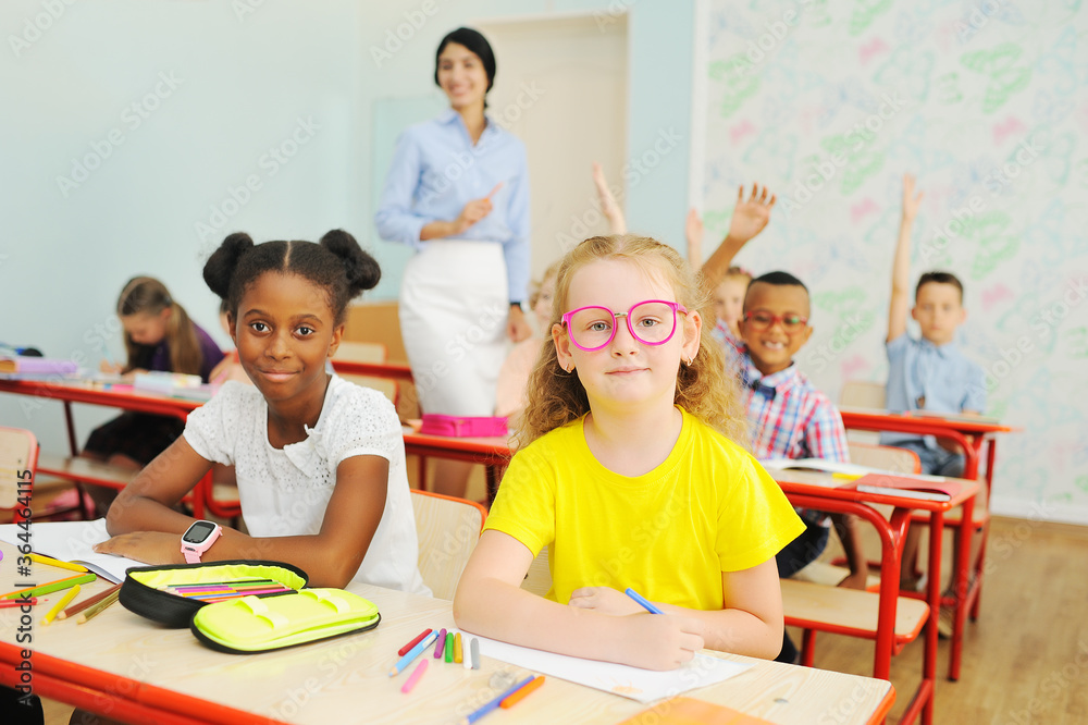 Fototapeta premium two children-an African-American girl and a Caucasian girl in pink glasses are smiling sitting at a Desk in a primary school against the background of a teacher 