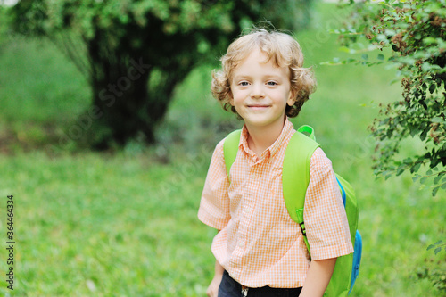 a small curly-haired schoolboy with a school bag smiles against the background of greenery and a Park. Back to school, education, knowledge day, vacation.