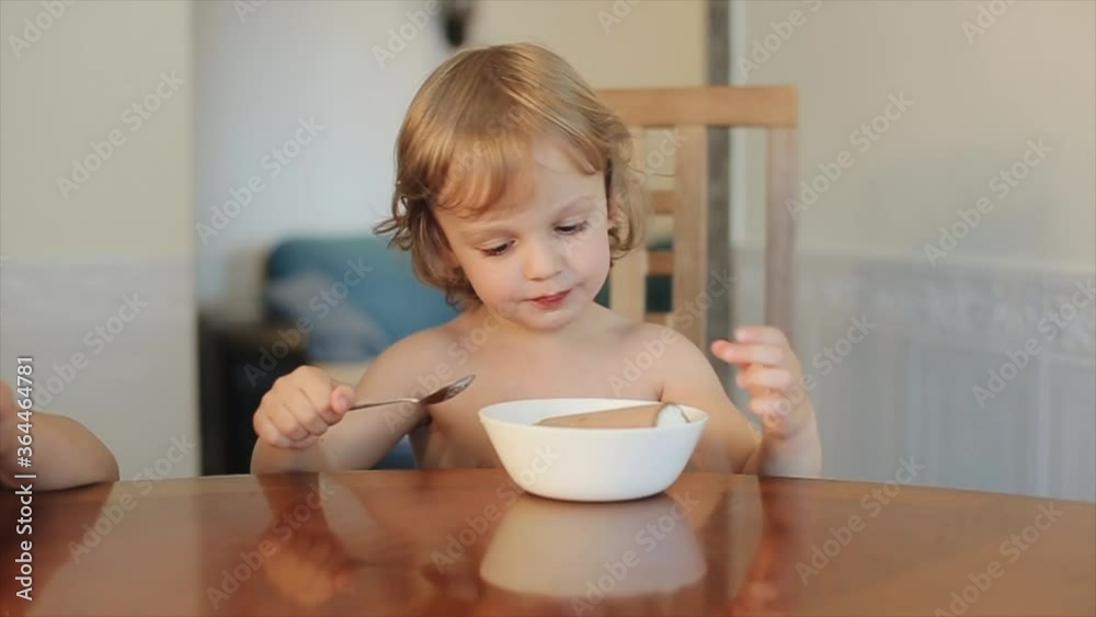 An enthusiastic little boy without clothes sits at the kitchen table and eating ice cream a spoon from a plate. Close-up An enthusiastic little boy without clothes sits at the kitchen table and eating ice cream a spoon from a plate. Close-up