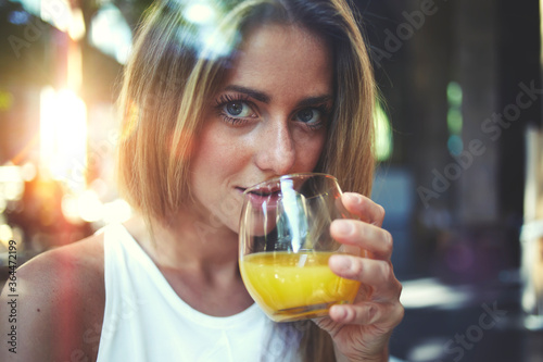 View though the window of a young gorgeous woman enjoying a natural fruit juice and looking at you, caucasian blonde hair female standing indoors and drinking freshly juiced beverage of orange