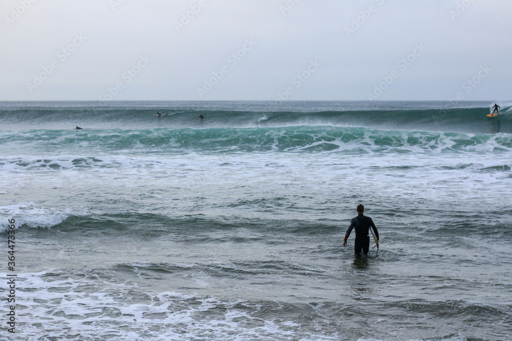 an unidentfiiable lone male surfer leading his surfboard through the waves off the beach on a rainy morning, as his friends surf in the background, Bell's beach, great ocean road, Victoria, Australia