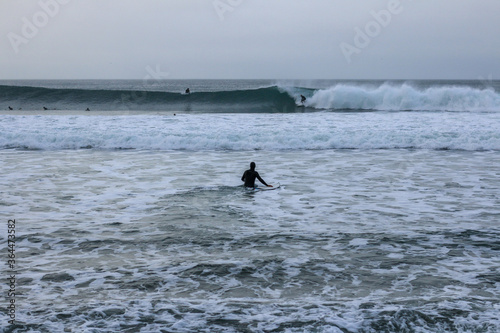 an unidentfiiable lone male surfer leading his surfboard through the waves off the beach on a rainy morning, as his friends surf in the background, Bell's beach, great ocean road, Victoria, Australia