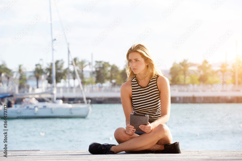 Fototapeta premium Young beautiful woman thinking about something after reading electronic book on her portable touch pad, dreaming female looking away while sitting with digital tablet on a wooden pier near sea port