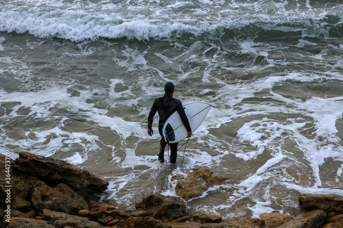 an unidentifiable lone male surfer carrying his surfboard over the rocks of the beach into the churning crashing waves on a rainy surf day, Bell's beach, great ocean road, Victoria, Australia