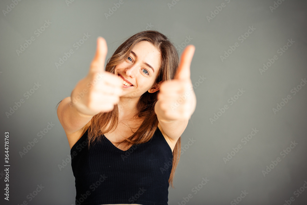 Fototapeta premium Girl in a black top with her hair shows thumb up. On a gray background. Without makeup, without retouching.