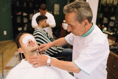 barber cutting the beard of an attractive young brown man with cotton wool in his eyes