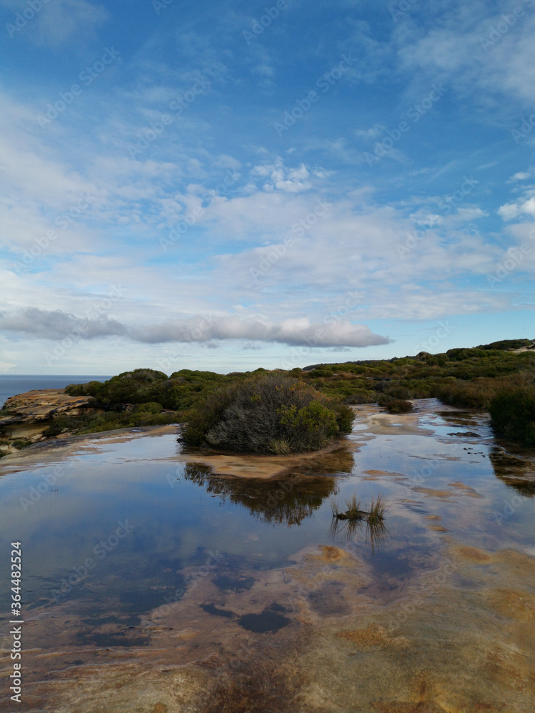 Beautiful coastal trail with colorful rock formations and reflections of blue sky on water puddles near Wattamolla Beach, Royal National Park, New South Wales, Australia