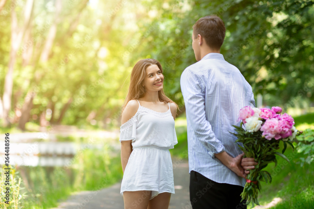 Fototapeta premium Young man congratulating girlfriend with birthday, hiding flower bouquet behind his back at park. Empty space
