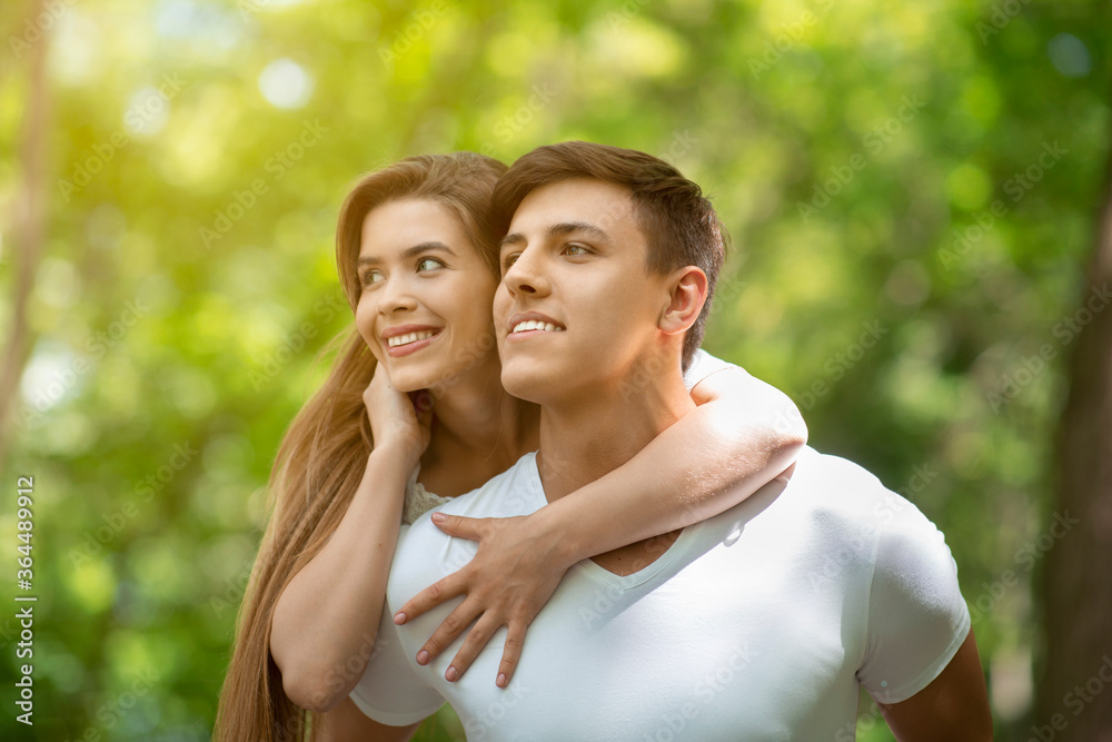 Attractive millennial guy carrying girlfriend on his back at green park Attractive millennial guy carrying girlfriend on his back at green park