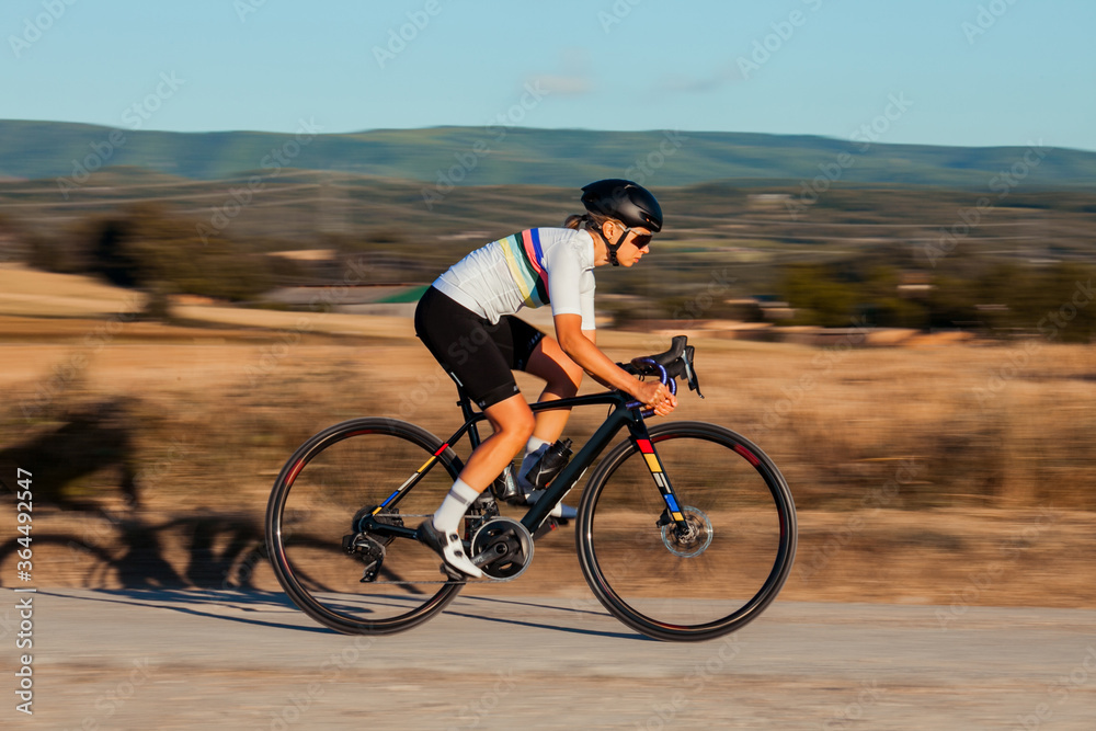 Professional beautiful blond young female athlete going down the hill very fast with her white cycling suit and on her black bicycle.