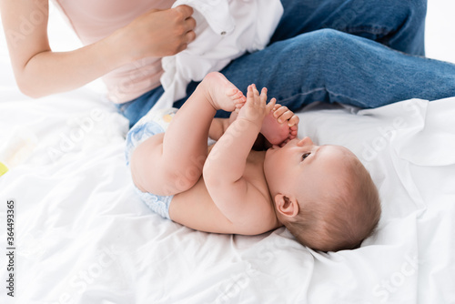 mother sitting on bed and holding baby romper near cute infant son in diaper