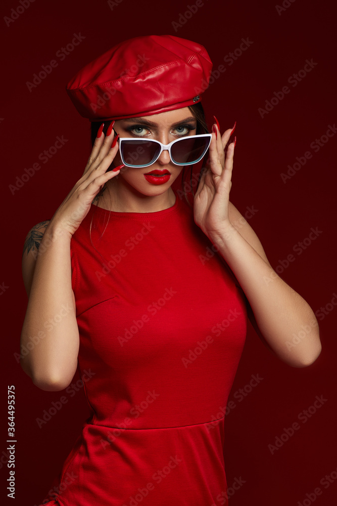 beauty stylish brunette young woman in trendy white sunglasses, hat and dress posing on red background. girl looking at the camera beauty stylish brunette young woman in trendy white sunglasses, hat and dress posing on red background. girl looking at the camera