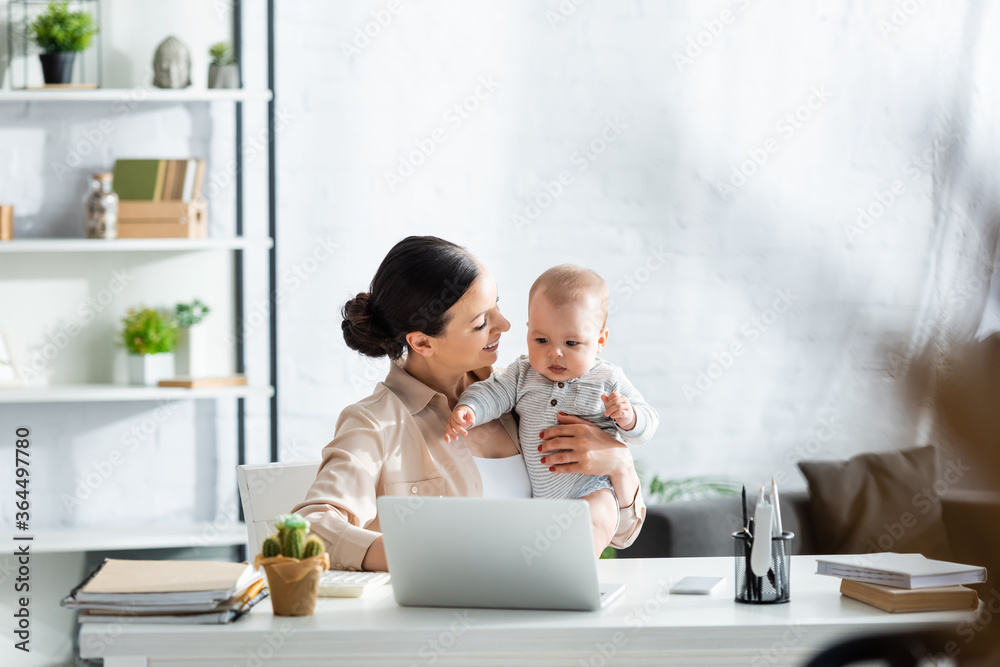 selective focus of happy mother looking at infant son near laptop on table selective focus of happy mother looking at infant son near laptop on table