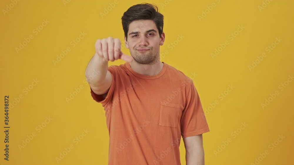 Displeased handsome man wearing in t-shirt showing thumb down gesture at the camera over yellow background Displeased handsome man wearing in t-shirt showing thumb down gesture at the camera over yellow background