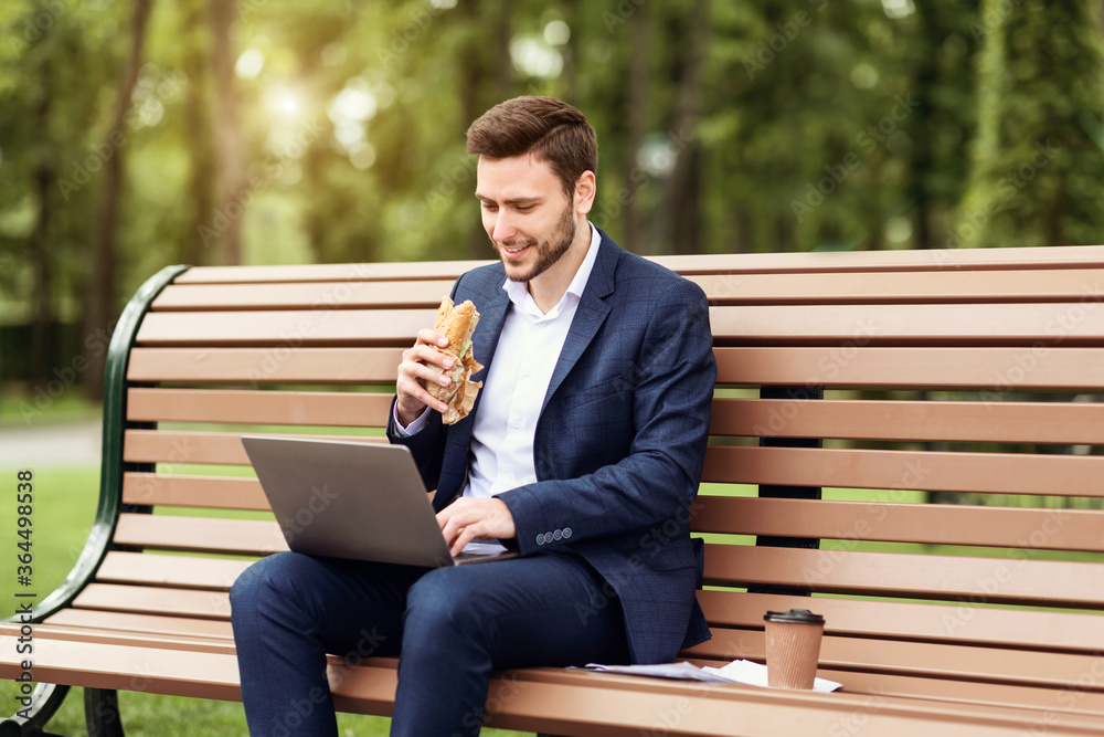 Fototapeta premium Millennial businessman eating sandwich and working on laptop during lunch break at park