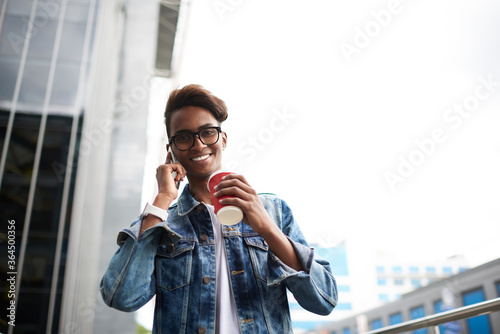 Below portrait of successful stylish blogger smiling at camera while drinking tasty coffee beverage and calling on smartphone.Happy hindu hipster guy in denim outfit talking on telephone on street