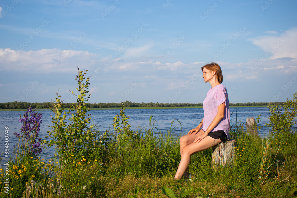The concept of walks alone. Young girl with short hair in purple T-shirt and shorts sits on river bank. Summer sunny evening. Copyspace. Cozy cute rural landscape.