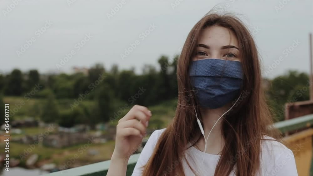 A romantic young girl in a medical mask walks on the bridge and listens to music with headphones. The camera changes focus. Slow motion. Close-up A romantic young girl in a medical mask walks on the bridge and listens to music with headphones. The camera changes focus. Slow motion. Close-up