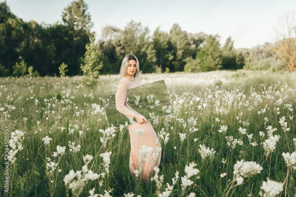 Young woman in beatiful dress walking with mirror in flowering meadow