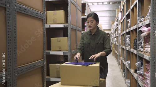 charming female warehouse worker pushing pallet truck with parcels and walking. asian woman employee stop and holding clipboard writing note while stock taking in stockroom standing with trolley