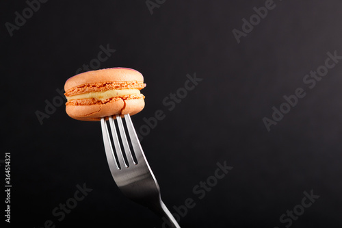 Single orange macaron or macaroon cake on a fork on black background. side view.