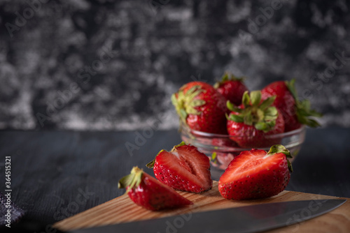 Fresas en un recipiente de cristal sobre una mesa de madera, con una tabla para cortar con fresas cortadas y un cuchillo al lado.